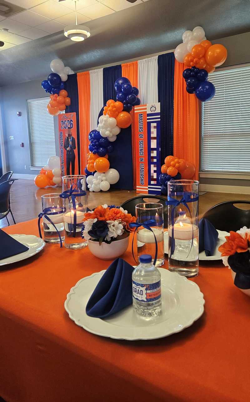 An orange dining table is set with white plates, blue napkins, and water bottles reading "Congrats GRAD." Centerpieces feature floating candles and flowers, backed by balloon columns and banners celebrating the Class of 2026.