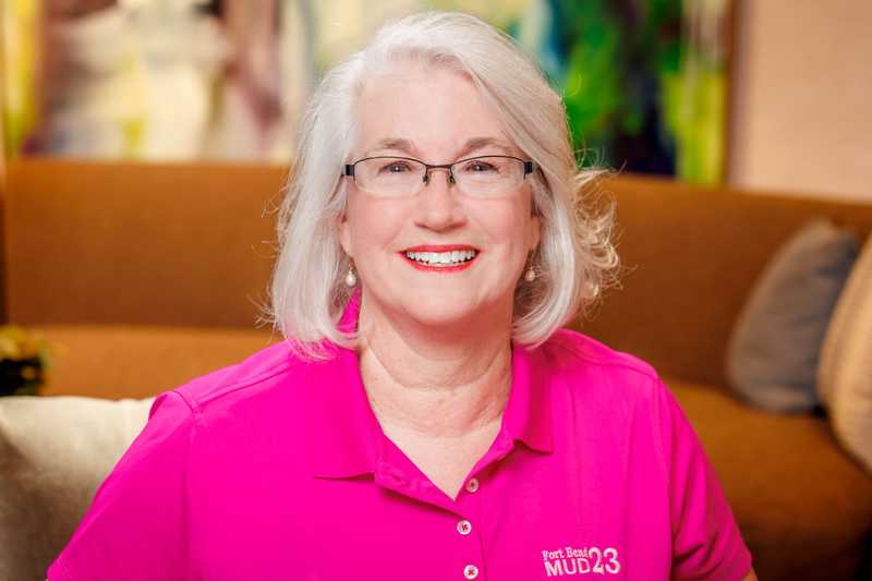 Professional headshot of a person wearing glasses and a bright pink Fort Bend MUD 23 polo shirt, seated indoors against a softly blurred background.