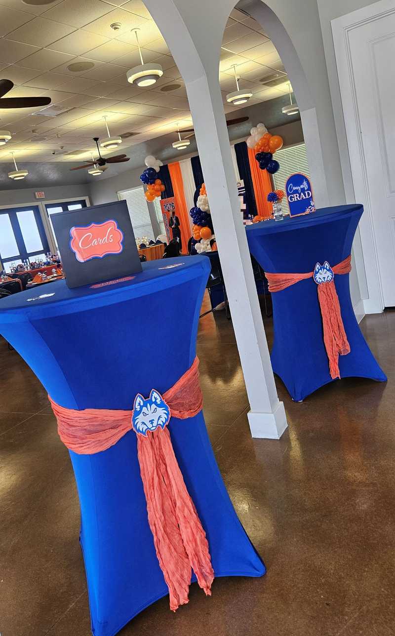 Two royal blue tables with coral sashes and a wolf logo stand near an archway. The left table holds a black "Cards" box, while the right displays a "Congrats GRAD" sign in front of orange and blue balloon decor.
